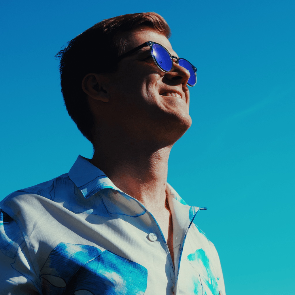 Ben Dava smiling in reflective blue sunglasses against a bright cyan sky